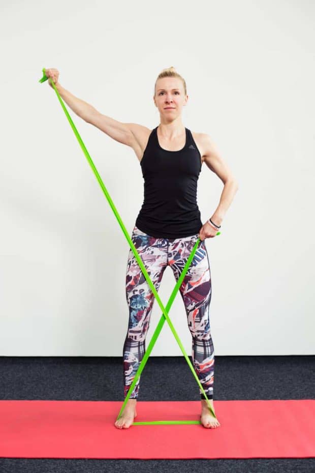 Frau auf roter Trainingsmatte steht frontal und macht Armheben diagonal mit dem Theraband