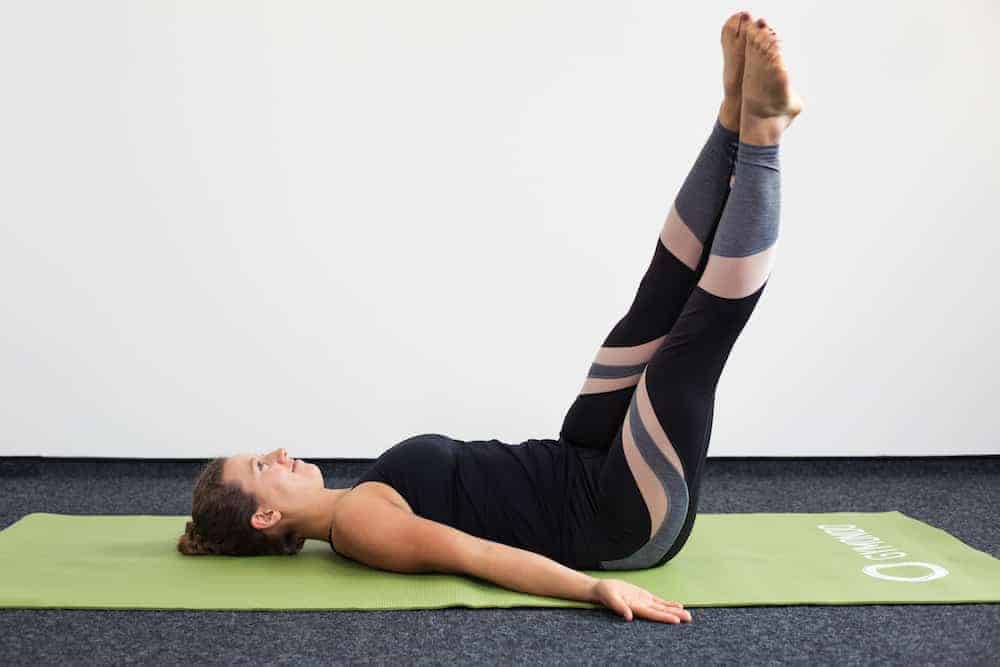 Junge Frau in Rückenlage auf einer hellgrünen Trainingsmatte macht Pilates Corkscrew