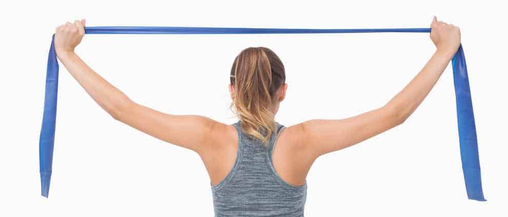 Junge Frau in hellgrauem Top von hinten zieht blaues Theraband auseinander und trainiert den Rücken - Rückenübungen Theraband