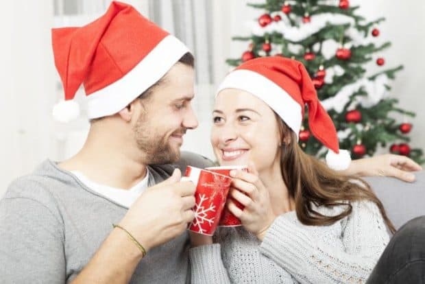 Ehepaar sitzt mit Weihnachtsmützen und Tassen auf dem Sofa vor einem Weihnachtsbaum