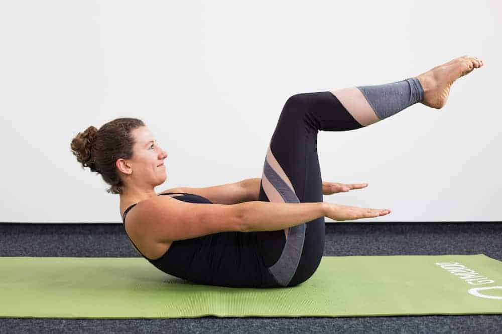 Junge Frau in Rückenlage mit angehobenen Beinen auf einer hellgrünen Trainingsmatte macht Pilates Hundred
