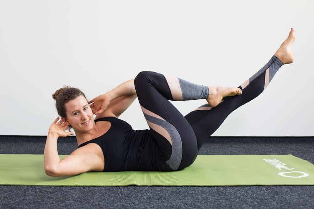Junge Frau in Rückenlage auf einer hellgrünen Trainingsmatte macht Pilates Criss Cross