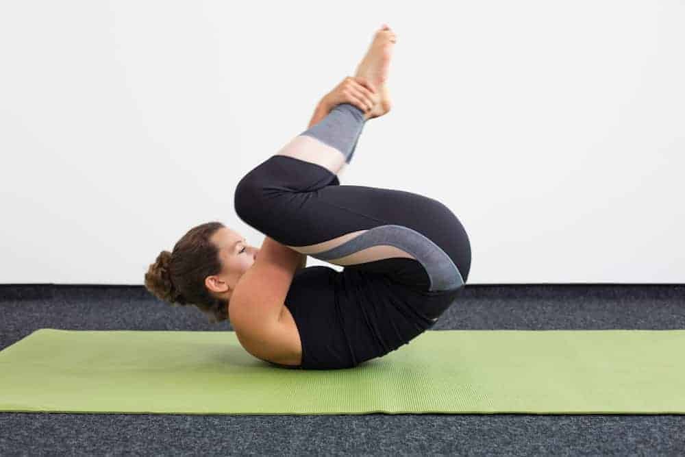 Junge Frau in Rückenlage auf einer hellgrünen Trainingsmatte macht Pilates Rolling Like a Ball