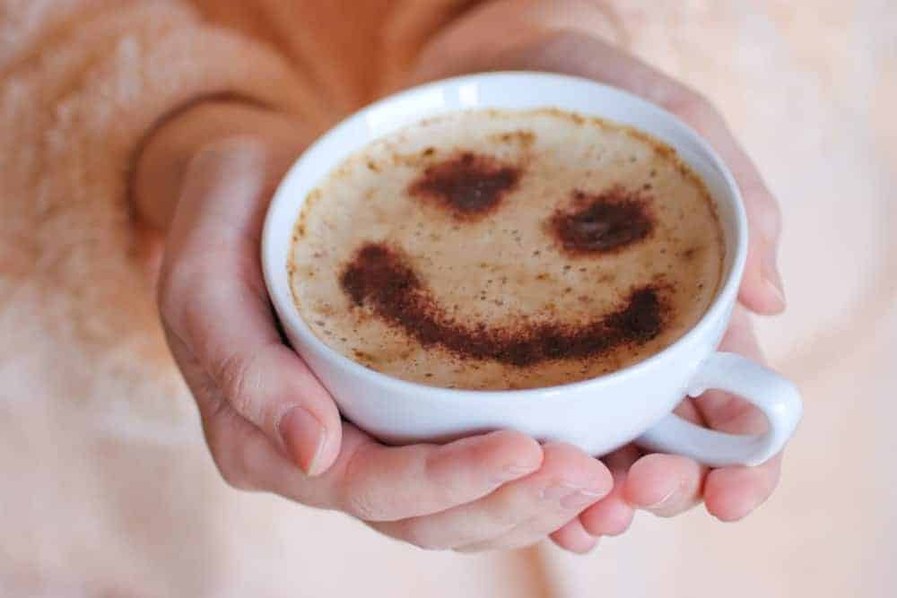 Hände einer Frau halten Kaffee-Tasse mit Milchkaffee auf dem Schaum ist ein Smiley mit Kakaopulver gestreut