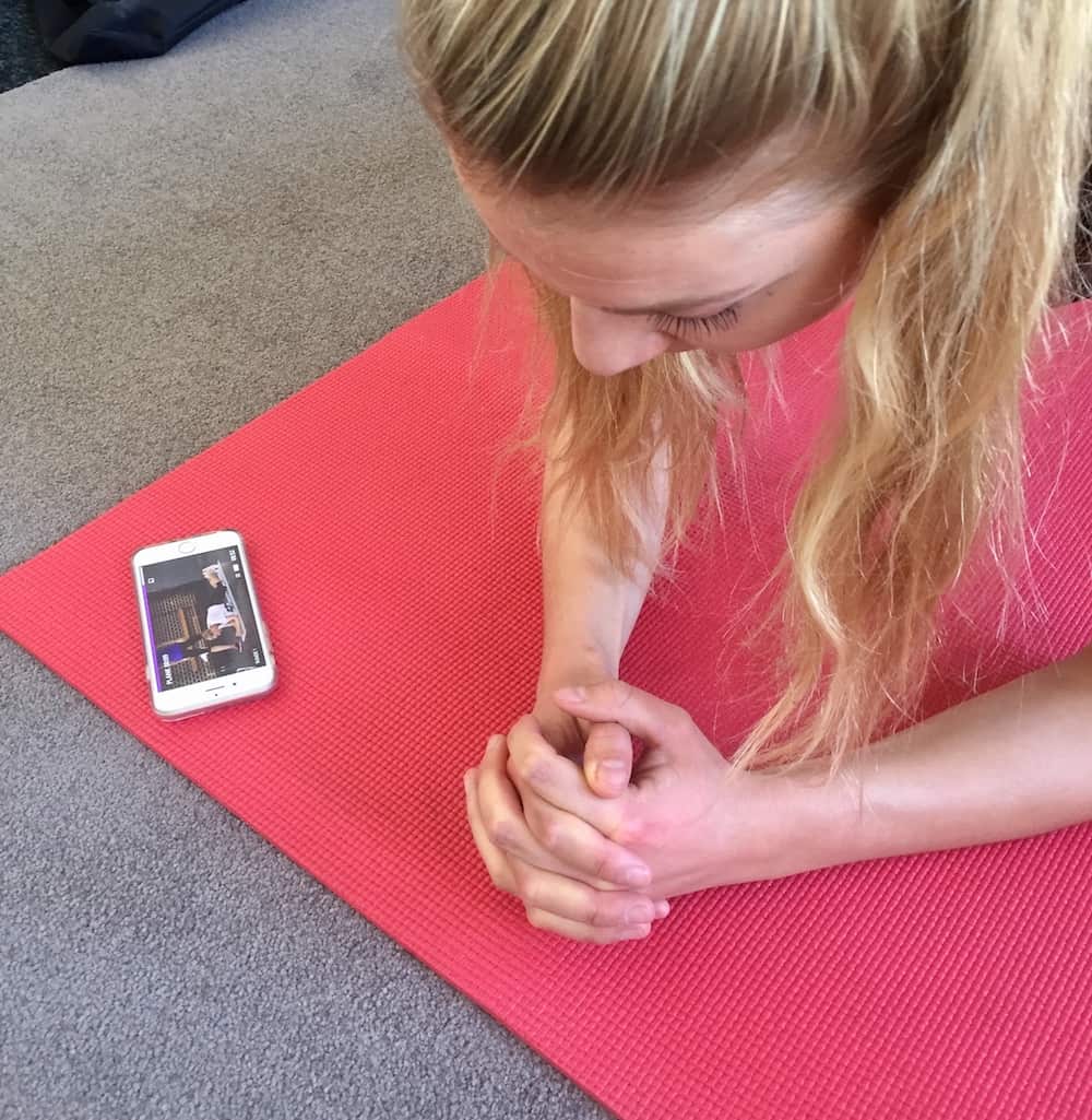 Junge Frau trainiert zu Hause auf roter Trainingsmatte. Weißes Smartphone liegt vor ihr. Gymondo Erfahrungen