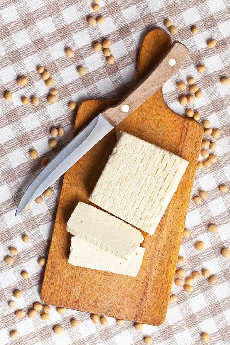 In Stücke geschnittener Tofu auf einem hölzernen Schneidebrett neben einem Messer umgeben von reifen Sojabohnen