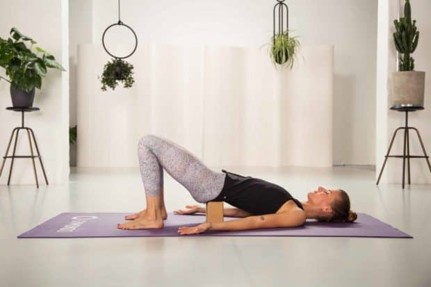 Junge Frau in Rückenlage mit angehobener Hüfte in hellem Raum auf lila Yogamatte mit geschlossenen Augen - Yin Yoga