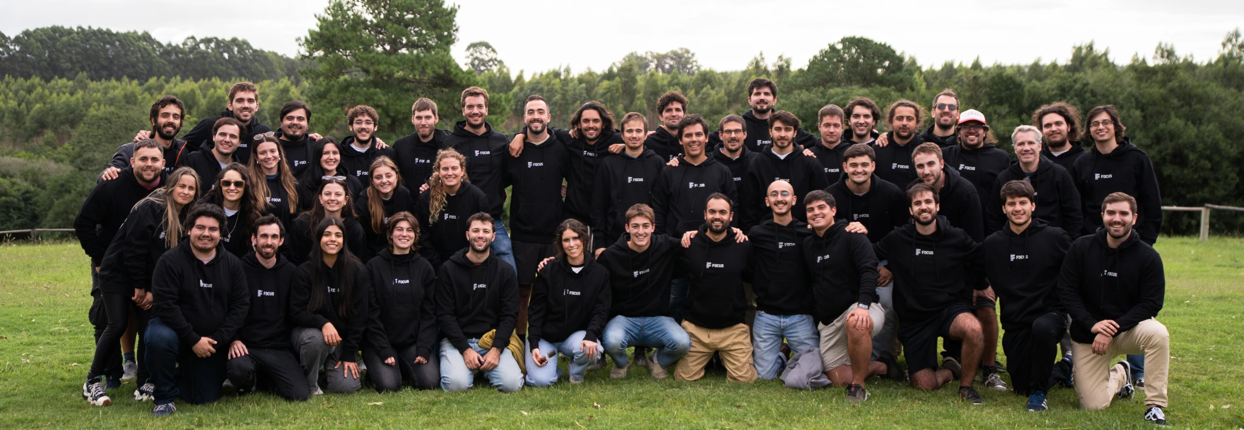 Large group of people wearing black hoodies with 'Focus' logo posing outdoors on grass with forest background.