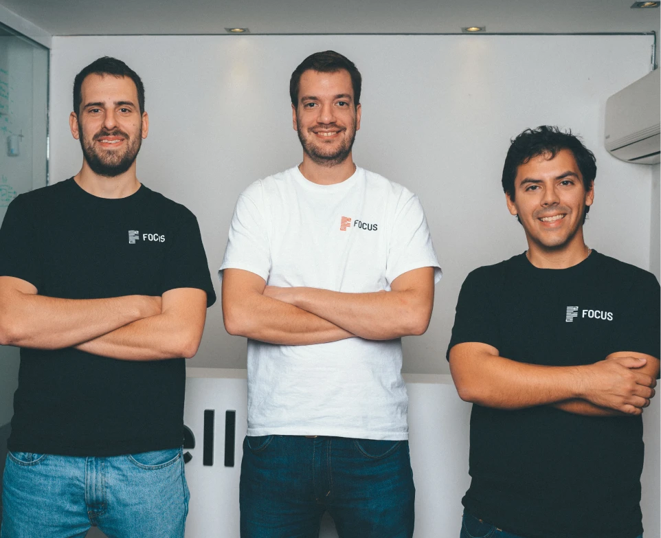 Three men standing indoors with arms crossed, two wearing black Focus t-shirts and one wearing a white Focus t-shirt, smiling at the camera.