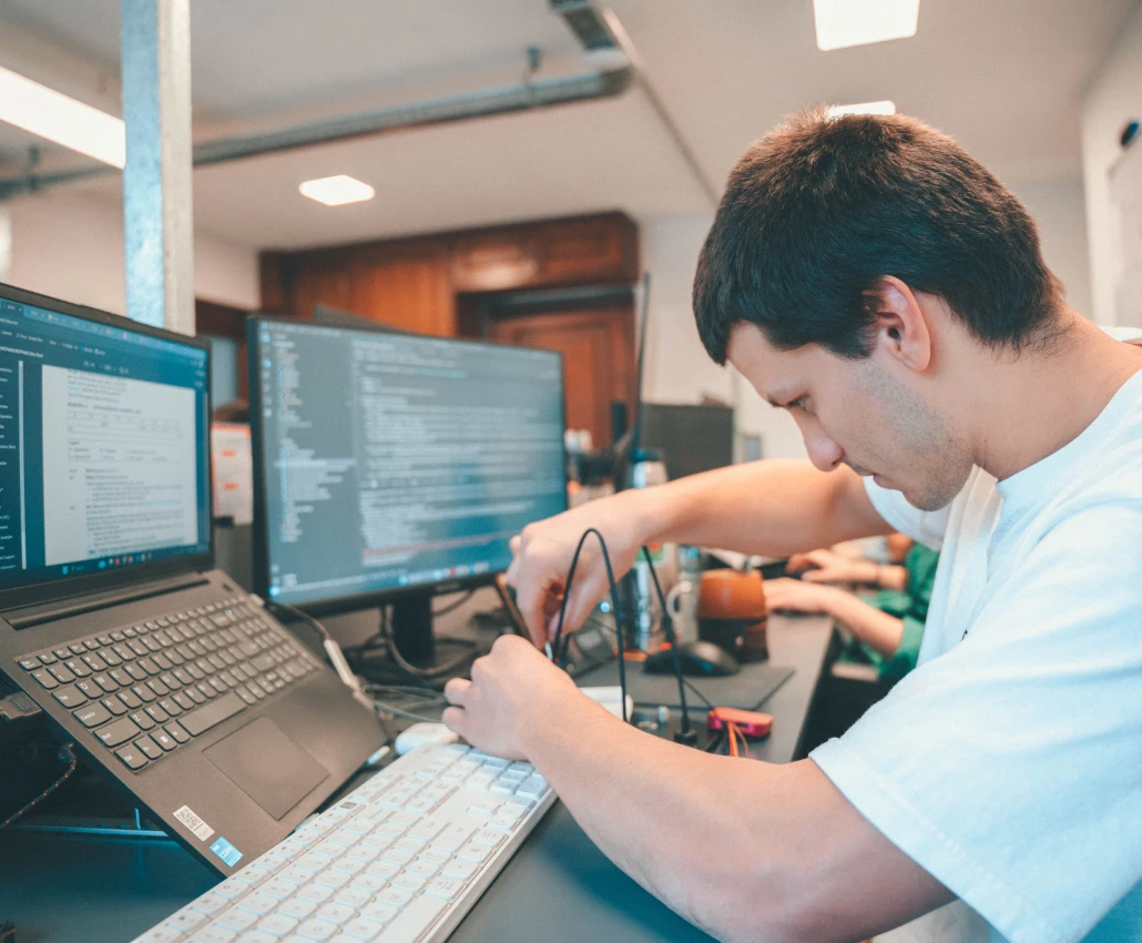 Young man intently working with electronic components and wires at a desk with two computer screens displaying code.