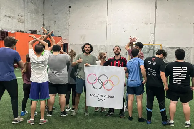 Group of people standing on indoor turf field, some facing away and others smiling holding a banner reading 'Focus Olympics 2023' with interlocking rings design.