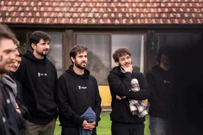 Group of casually dressed men standing outdoors in front of a building with a tiled roof, some wearing black hoodies and holding items.