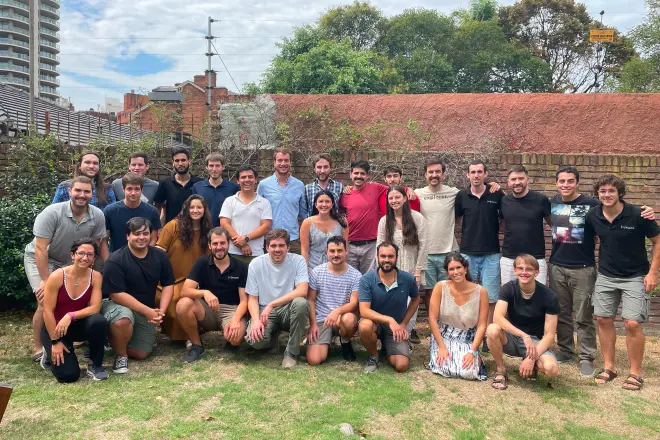 Group of 24 people posing outdoors on grass in front of a brick wall with trees and buildings in the background.