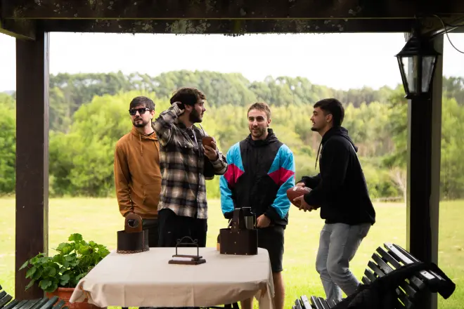 Four young men standing around a table with objects on it in an outdoor covered area with a grassy field and trees in the background.
