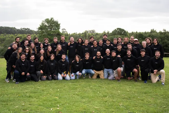 Group of 40 people wearing black sweatshirts posing together on green grass with trees in the background under a cloudy sky.