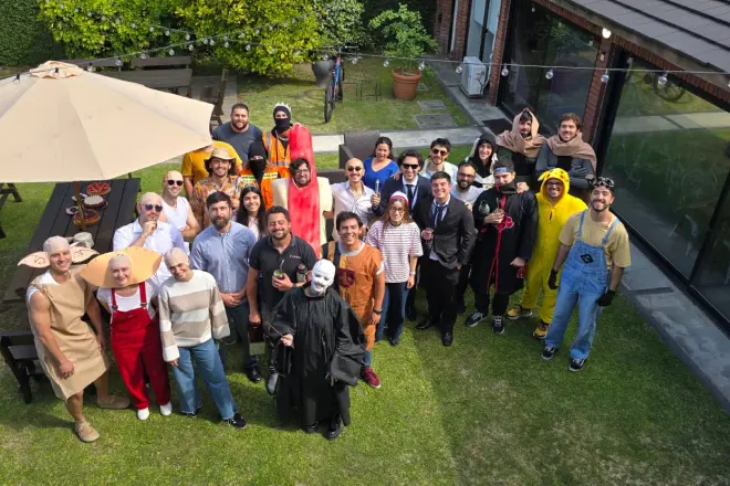 Group of people posing outdoors in various costumes including a hotdog, Pikachu, and others under a large umbrella near a picnic table.