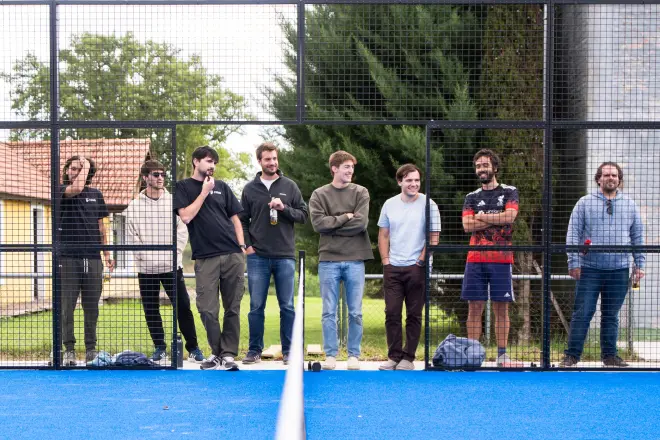 Eight casually dressed men standing behind a wire fence near a blue sports court with trees and buildings in the background.