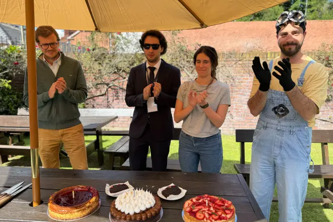 Four people standing outdoors under an umbrella, clapping near a table with cakes and dessert plates.