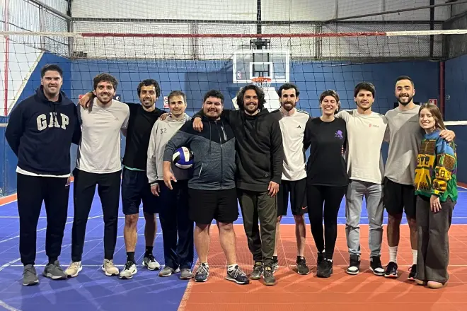 Group of ten people standing together on an indoor volleyball court with a volleyball net behind them, one person holding a volleyball.