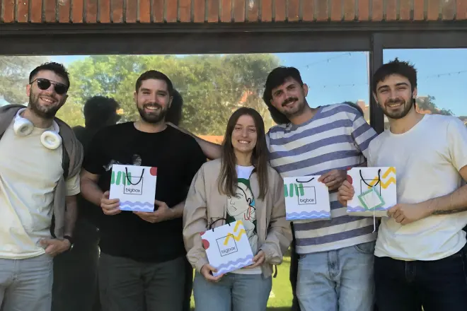 Group of five smiling people standing outdoors holding branded gift bags in front of a glass window.