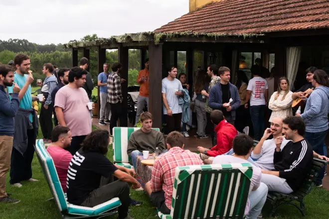 Group of people socializing outdoors on a grassy lawn and porch of a house with striped patio chairs and casual clothing.