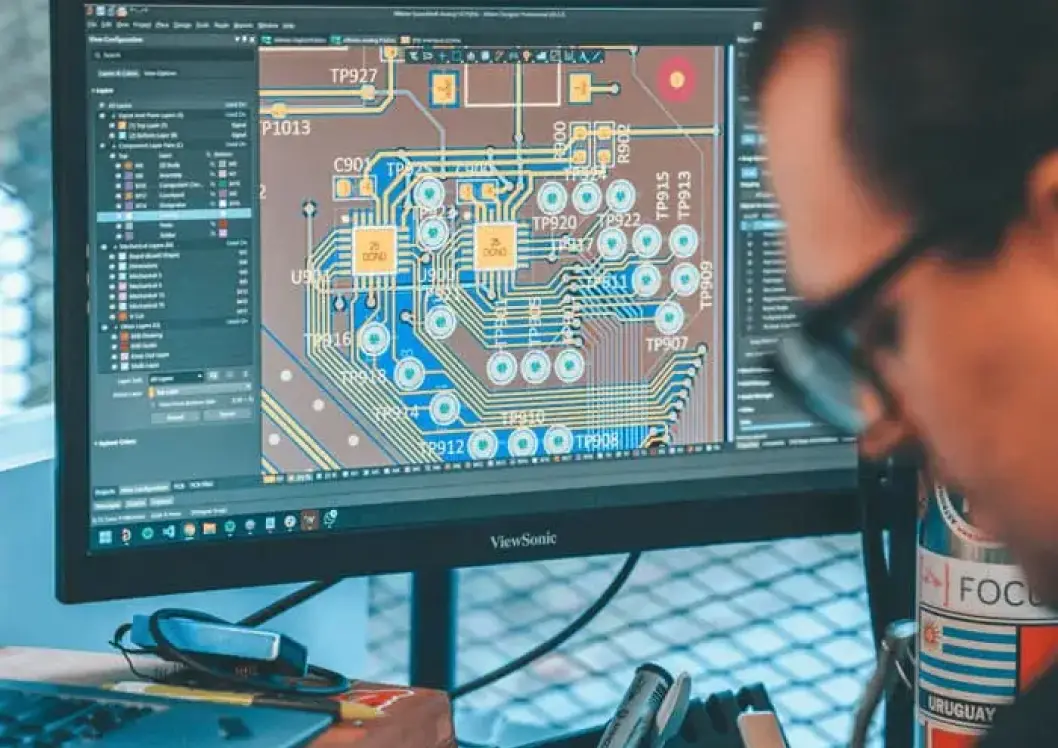 Close-up of a computer monitor displaying a detailed electronic circuit board design with various connections and components, with a person wearing glasses looking at the screen.