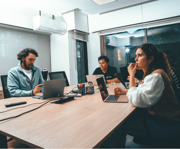 Three people sitting around a wooden table in an office room with laptops, appearing focused and engaged in discussion, with one person gesturing and another person resting their chin on their hand.
