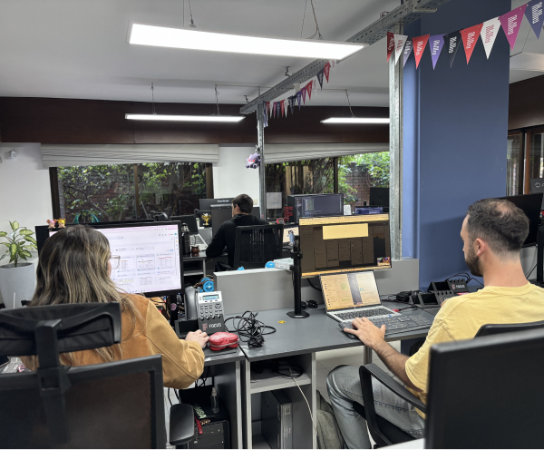 Office scene with two people seated at desks working on computers, one looking at a large monitor and the other using a laptop, with a third person visible in the background also working at a computer near large windows showing outdoor greenery.