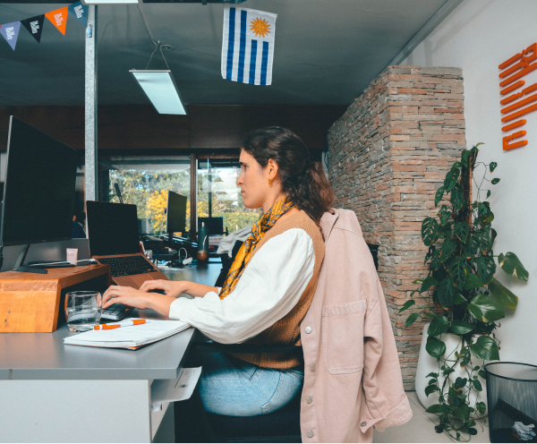 A woman with a beige jacket draped over her chair working on a laptop at a desk in an office with multiple monitors and a plant beside a stone wall.