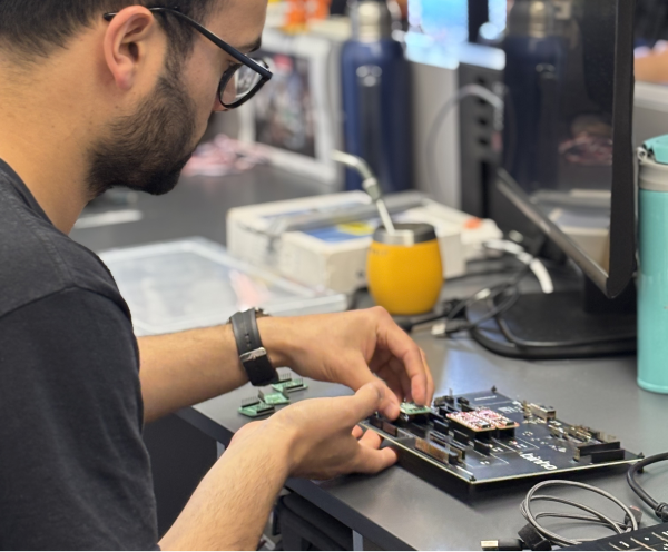Man with glasses assembling or inspecting a circuit board at a desk with computer monitor, cables, and a yellow cup in the background.