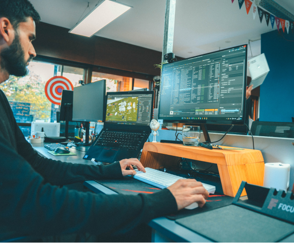 Man with beard working at desk using a keyboard and mouse, with a laptop and a large monitor displaying data and graphs in a brightly lit office space.