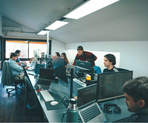 Office workspace with several people working on computers arranged in a row, featuring monitors, laptops, headphones, and other office equipment under ceiling lights and near a large window.