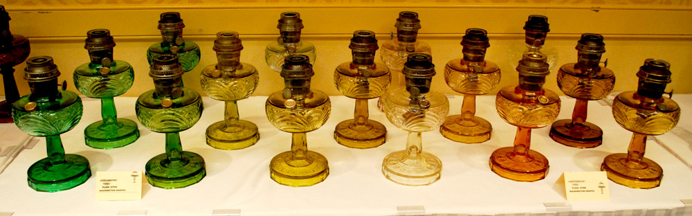 Rows of vintage glass oil lamps in green, yellow, clear, and amber colors displayed on a white tablecloth.