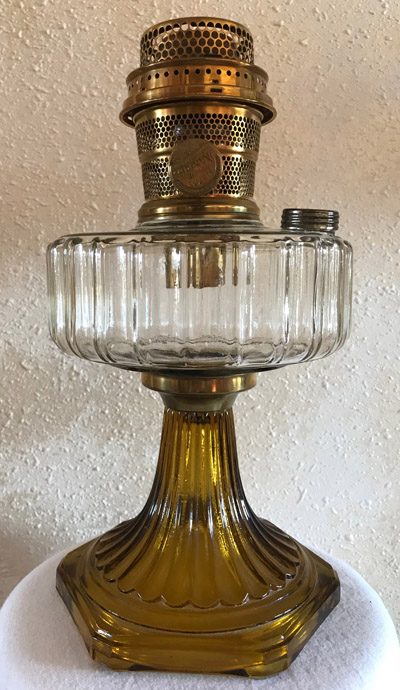 Vintage amber glass oil lamp with clear ribbed glass fuel reservoir and brass burner on textured wall.