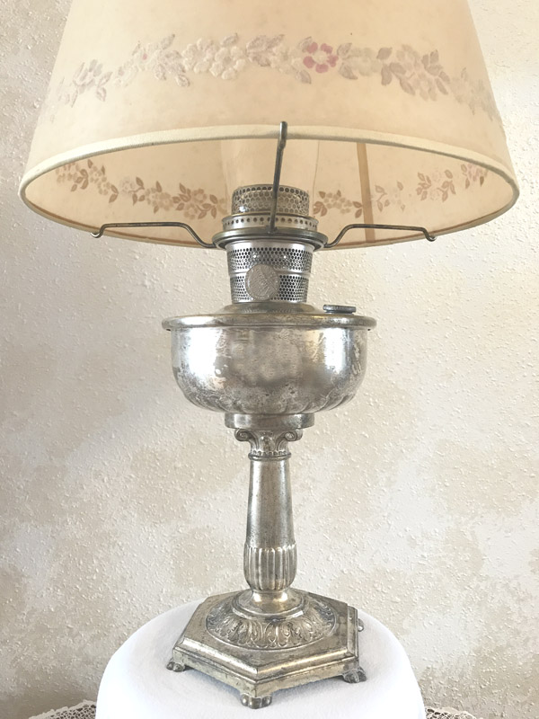 Antique silver oil lamp with beige floral lampshade on a white surface against a beige wall.