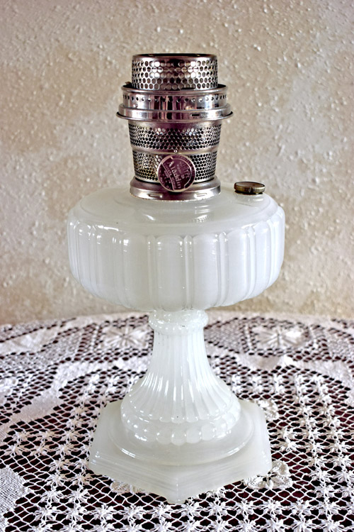 White vintage milk glass oil lamp with ornate base and metal burner on lace tablecloth.
