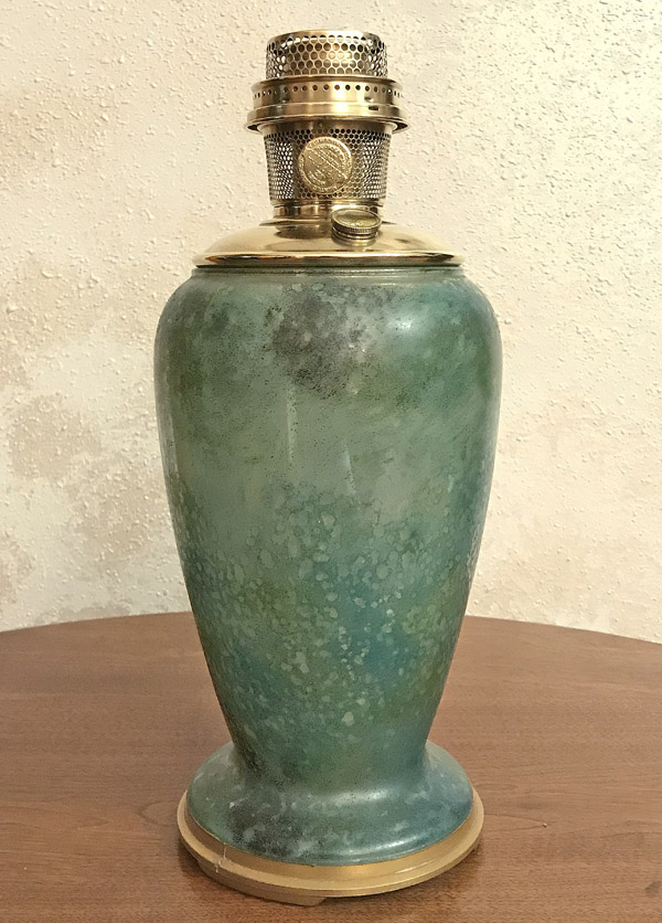 Greenish-blue variegated glass lamp base with brass burner on a wooden surface.