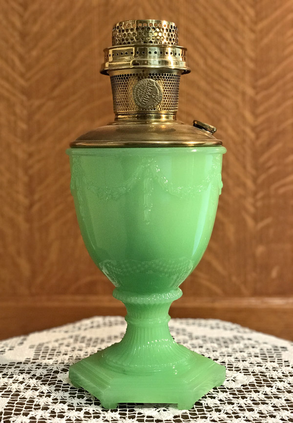 Ornate green glass oil lamp with brass burner on a lace-covered surface.