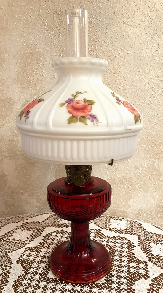 Vintage oil lamp with white floral glass shade and deep red glass base on lace tablecloth.