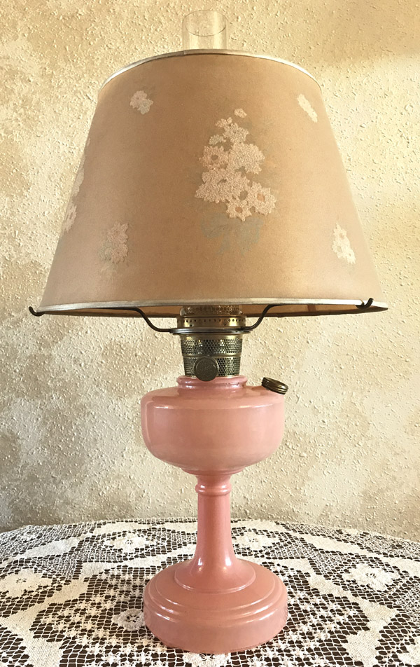 Vintage pink oil lamp with beige floral lampshade on a lace tablecloth against a textured wall.