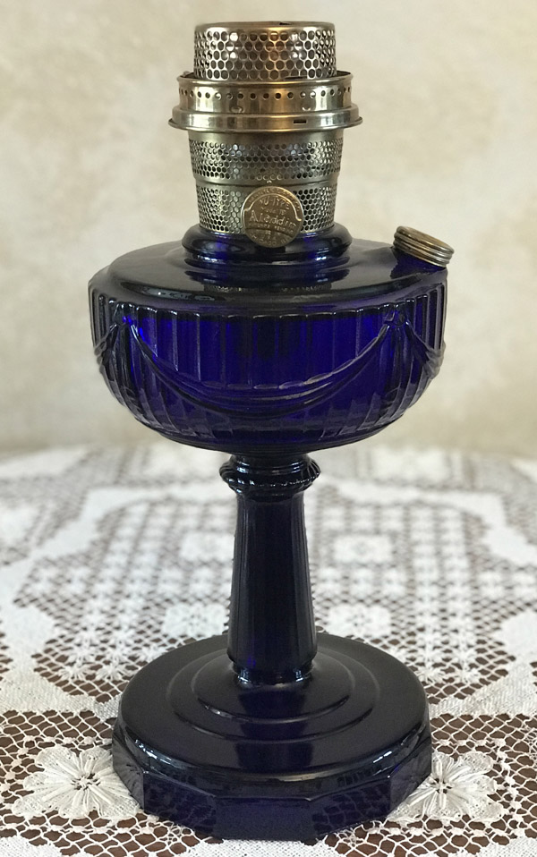 Antique cobalt blue glass oil lamp with metal burner on a white lace tablecloth.