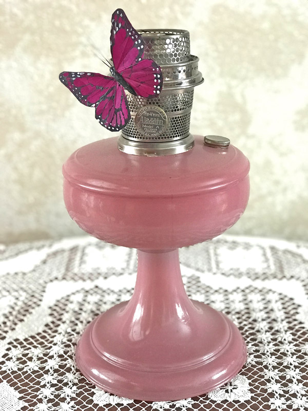 Pink vintage Aladdin oil lamp with metal burner, adorned with a purple butterfly decoration.