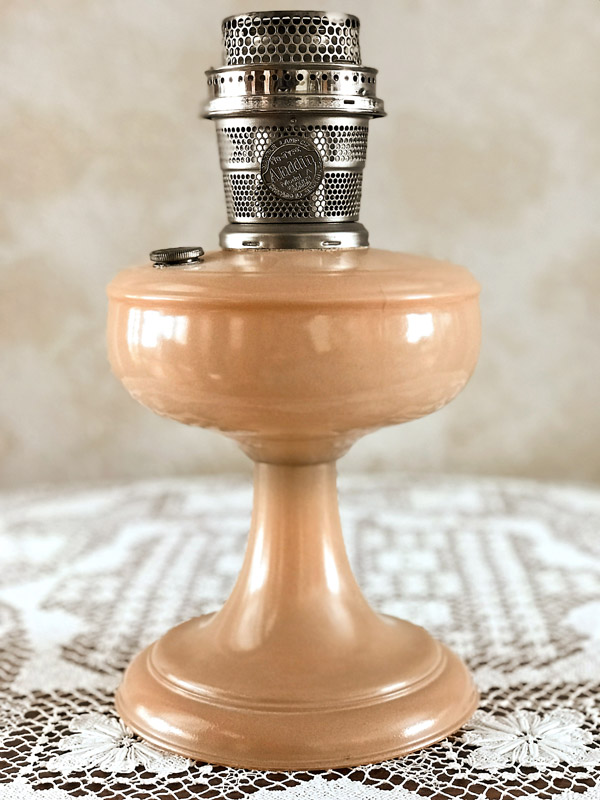 Vintage peach-colored Aladdin kerosene lamp on lace tablecloth with a beige background.