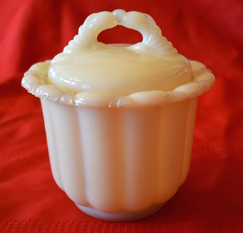 Cream-colored vintage glass candy dish with lid and ornate handle on red fabric background.