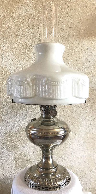 Vintage silver oil lamp with ornate white glass shade and clear chimney on textured wall background.