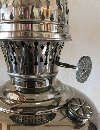 Close-up of a shiny metal vintage kerosene lamp burner with adjustable wick knob and decorative vents.
