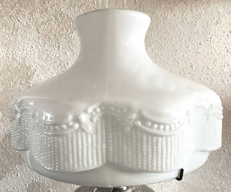 White opaque glass lamp shade with raised bead and swag decorations on a textured wall background.