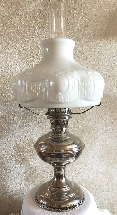 Vintage kerosene lamp with ornate white glass shade and polished silver base against textured wall.