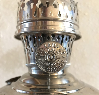 Close-up of a metal antique oil lamp valve knob with embossed text and decorative perforations on the chimney.
