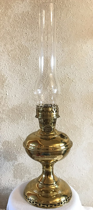 Polished brass oil lamp with glass chimney on a white cloth against a textured wall.